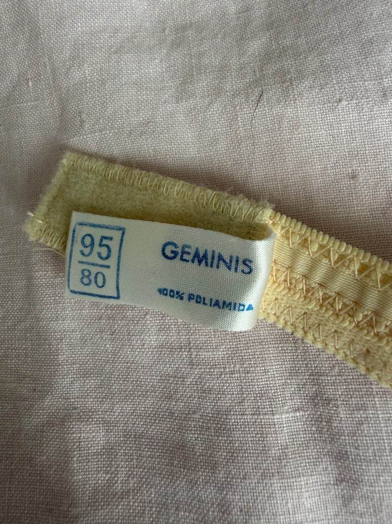 May include: Close-up of a pale yellow bra strap with a label. The label reads "GEMINIS" and "100% POLIAMIDA" with size markings "95" and "80". The strap is on a pink fabric background.