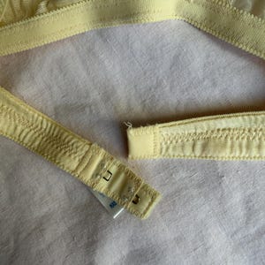 May include: Close-up of a pale yellow bra with delicate floral lace detailing. The bra features adjustable straps and a hook-and-eye closure. The fabric appears soft and lightweight, with a subtle sheen.