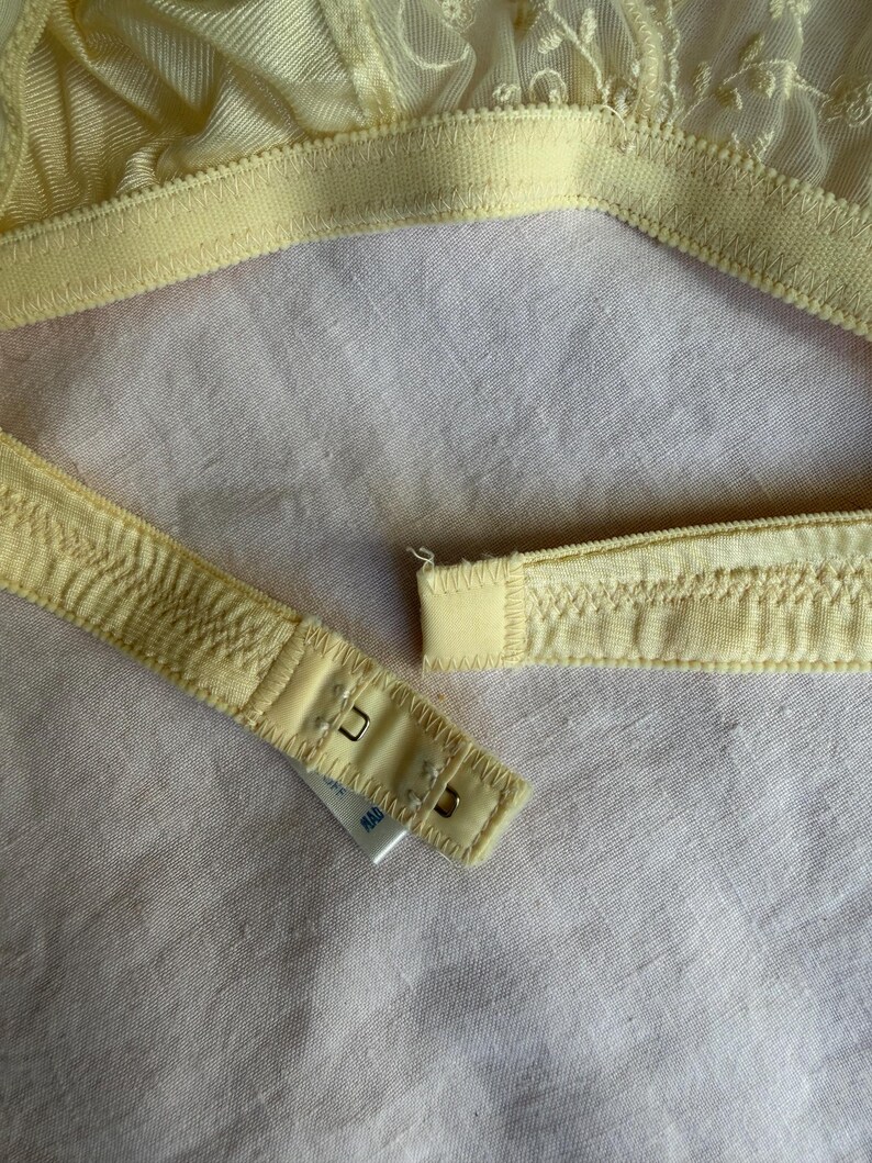May include: Close-up of a pale yellow bra with delicate floral lace detailing. The bra features adjustable straps and a hook-and-eye closure. The fabric appears soft and lightweight, with a subtle sheen.