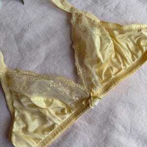 May include: A pale yellow bra with delicate lace detailing. The bra features a soft, satin-like fabric and floral lace accents along the cups and straps. A small bow adorns the center. The bra is designed with a classic triangle shape.