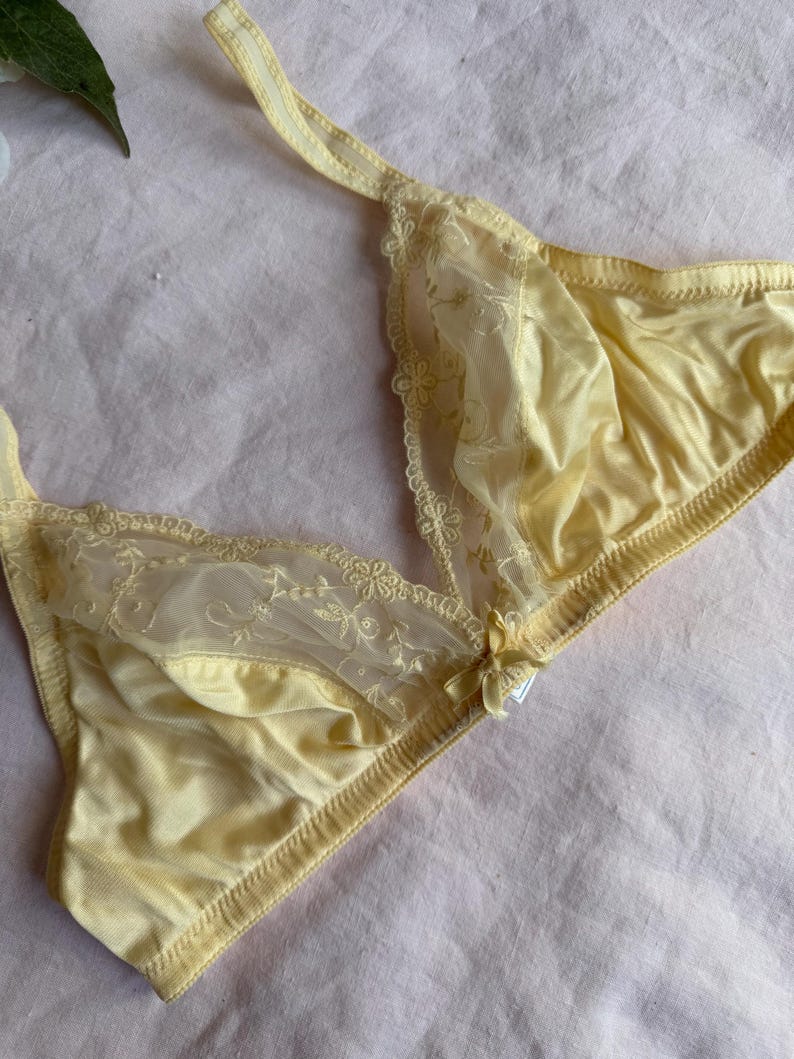 May include: A pale yellow bra with delicate lace detailing. The bra features a soft, satin-like fabric and floral lace accents along the cups and straps. A small bow adorns the center. The bra is designed with a classic triangle shape.