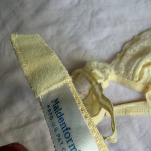 May include: Close-up of a pale yellow Maidenform bra. The bra features delicate lace detailing and adjustable straps. The tag reads "Maidenform *REG. U.S. PAT. OFF MADE IN SPAIN".