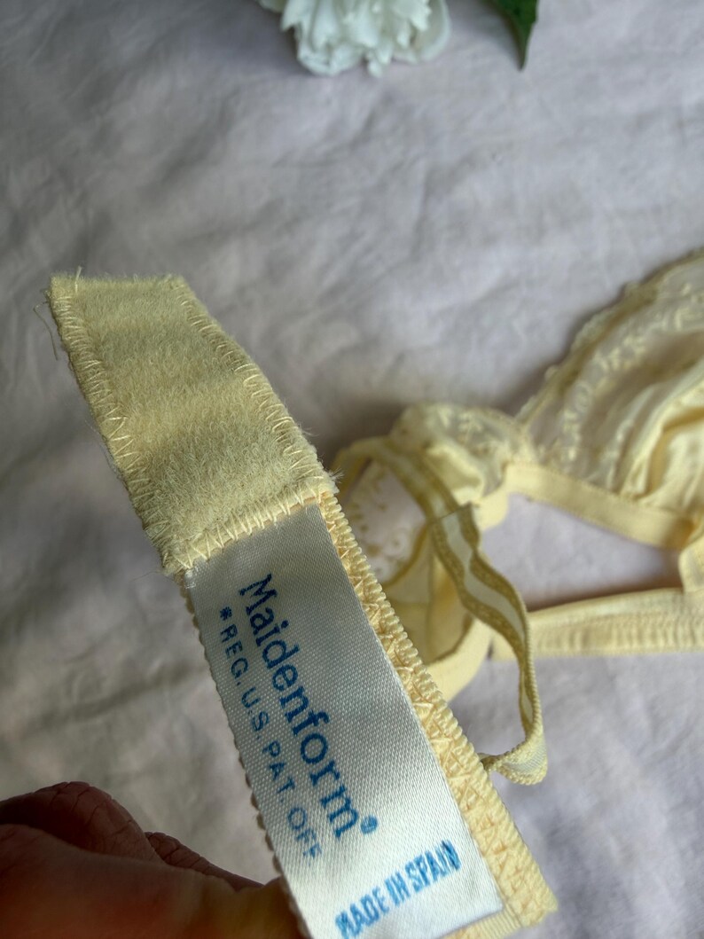 May include: Close-up of a pale yellow Maidenform bra. The bra features delicate lace detailing and adjustable straps. The tag reads "Maidenform *REG. U.S. PAT. OFF MADE IN SPAIN".