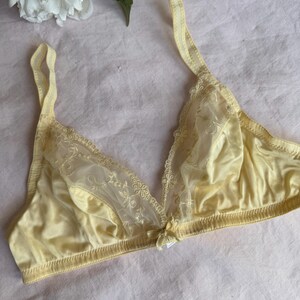 May include: A pale yellow bra with delicate lace detailing on the cups and straps. The bra features a small bow at the center and is made of a soft, silky material. The background is a light pink fabric.