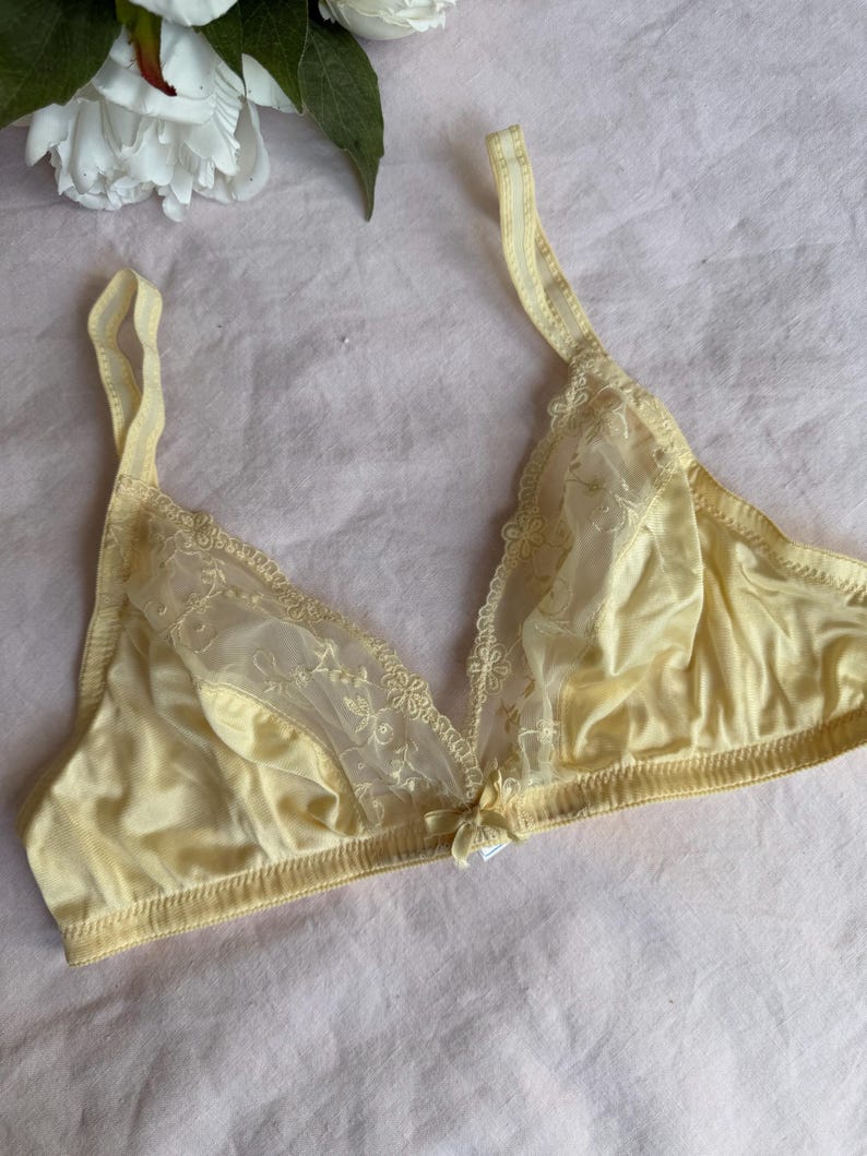 May include: A pale yellow bra with delicate lace detailing on the cups and straps. The bra features a small bow at the center and is made of a soft, silky material. The background is a light pink fabric.