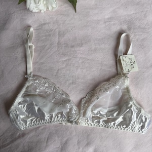 May include: A white satin bra with delicate lace detailing along the edges and straps. The bra features a sheer panel and a tag that reads "Little K". The bra is displayed on a pink surface.