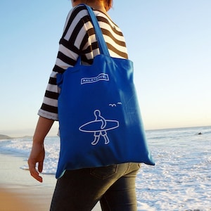 May include: A blue tote bag with a white surfer graphic and the text "BALATATravel" on the front. The bag is being carried by a person wearing a striped shirt and jeans.