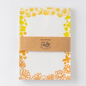 Può includere: Un blocco note bianco con un bordo floreale giallo e arancione. Il blocco note ha una fascia di carta marrone con il testo "NOTEPAD Lala Balaclara.com".