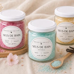 Peut inclure: Trois pots en verre de sels de bain avec des couvercles blancs. Les pots contiennent des sels roses, bleus et jaunes. Chaque pot porte une étiquette avec le texte "SELS DE BAIN NATURELS". Une cuillère en bois et des fleurs sont également présentes.