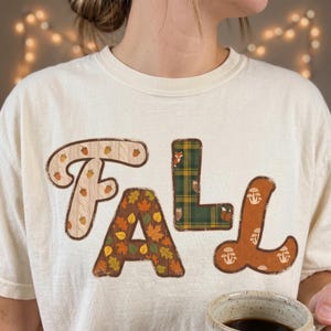 Puede incluir: Una camiseta de cuello redondo color crema con la palabra FALL en letras grandes y estilizadas. Cada letra tiene un patrón único: la F tiene bellotas sobre una textura de punto, la A hojas de otoño, la L animales del bosque sobre cuadros y la L setas.