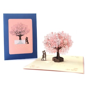 May include: A 3D pop-up card depicting a pink cherry blossom tree with a couple standing beneath it. The card is closed and shows a pink background with the couple and tree in silhouette.