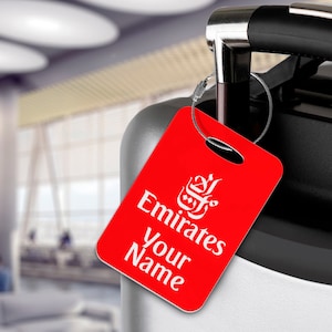 Könnte beinhalten: Roter Gepäckanhänger mit weißem Text, der "Emirates" und "Your Name" lautet. Der Anhänger zeigt das Emirates-Logo in Weiß.