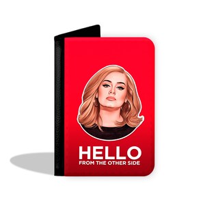 Puede incluir: Cartera de pasaporte roja con borde negro. La parte delantera presenta una ilustración de dibujos animados de Adele con el texto "HELLO FROM THE OTHER SIDE".