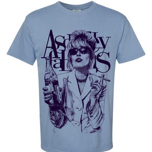 May include: Light blue t-shirt featuring a purple graphic of a woman in sunglasses holding a bottle and cigarette. The text "Absolutely Fabulous" is printed above the image in a stylized font.