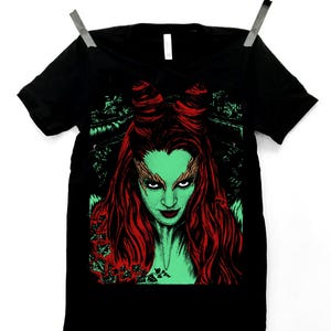 Pode incluir: Camiseta preta com um gráfico de uma mulher com pele verde e cabelo vermelho. O design inclui elementos botânicos vermelhos e verdes. A camiseta está pendurada em um fundo branco.