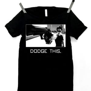 May include: Black t-shirt with a graphic of a person holding a gun, with the text "DODGE THIS." The image is in black and white. The t-shirt is hanging from a clothesline.