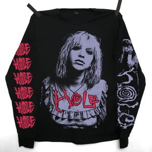 May include: Black long-sleeved shirt with a portrait of a person with purple hair and the word "HOLE" in red. The sleeves display the word "HOLE" and a spiral design in red and white.