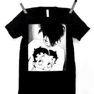 May include: Black t-shirt with a white graphic print of a cartoon character with large eyes and a man with long hair.