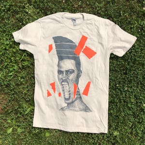 Könnte beinhalten: Cremefarbenes T-Shirt mit einem grafischen Porträt einer Person mit offenem Mund und einer Hochsteckfrisur. Die Grafik wird durch orangefarbene geometrische Formen akzentuiert. Das T-Shirt liegt auf grünem Gras.