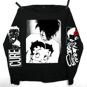 Pode incluir: Camiseta preta de manga comprida com um gráfico branco de um homem com um rosto de mulher sobreposto em seu peito. O rosto da mulher é um desenho animado de Betty Boop. As mangas têm a palavra "CURE" impressa em branco.