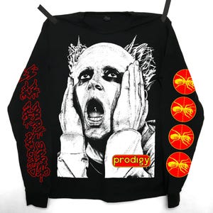 Peut inclure: T-shirt noir à manches longues avec un graphisme en noir et blanc d'une personne les mains sur le visage et la bouche ouverte. Le mot "prodigy" est dans un rectangle rouge et jaune. Du texte rouge est sur la manche gauche et des graphiques de fourmis rouges sur la manche droite.