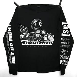 Könnte beinhalten: Schwarzes Langarmshirt mit einem Roboter-Motiv und dem Text "Toonami Deep Space Bass". Der linke Ärmel trägt "FALL DOWN SEVEN TIL GET UP EIGHT" und der rechte Ärmel "[as]" und Anime-Titel. Das Shirt hängt.