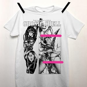 Puede incluir: Camiseta blanca con un gráfico en blanco y negro de Ghost in the Shell. El gráfico presenta tres paneles con ilustraciones de un personaje en un entorno futurista. El texto en la camiseta dice "People love machines in 2029 A.D." y "Ghost in the Shell."