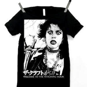 Könnte beinhalten: Schwarzes T-Shirt mit einer schwarz-weißen Grafik einer Frau mit großen Augen und einem schockierten Gesichtsausdruck. Die Grafik enthält japanischen Text und den Satz "WELCOME TO THE WITCHING HOUR". Das T-Shirt hängt vor einem weißen Hintergrund.