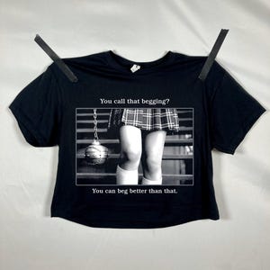 May include: Black crop top with a grayscale image of a wrecking ball and legs wearing a plaid skirt and knee socks. Text on the shirt reads, "You call that begging? You can beg better than that."