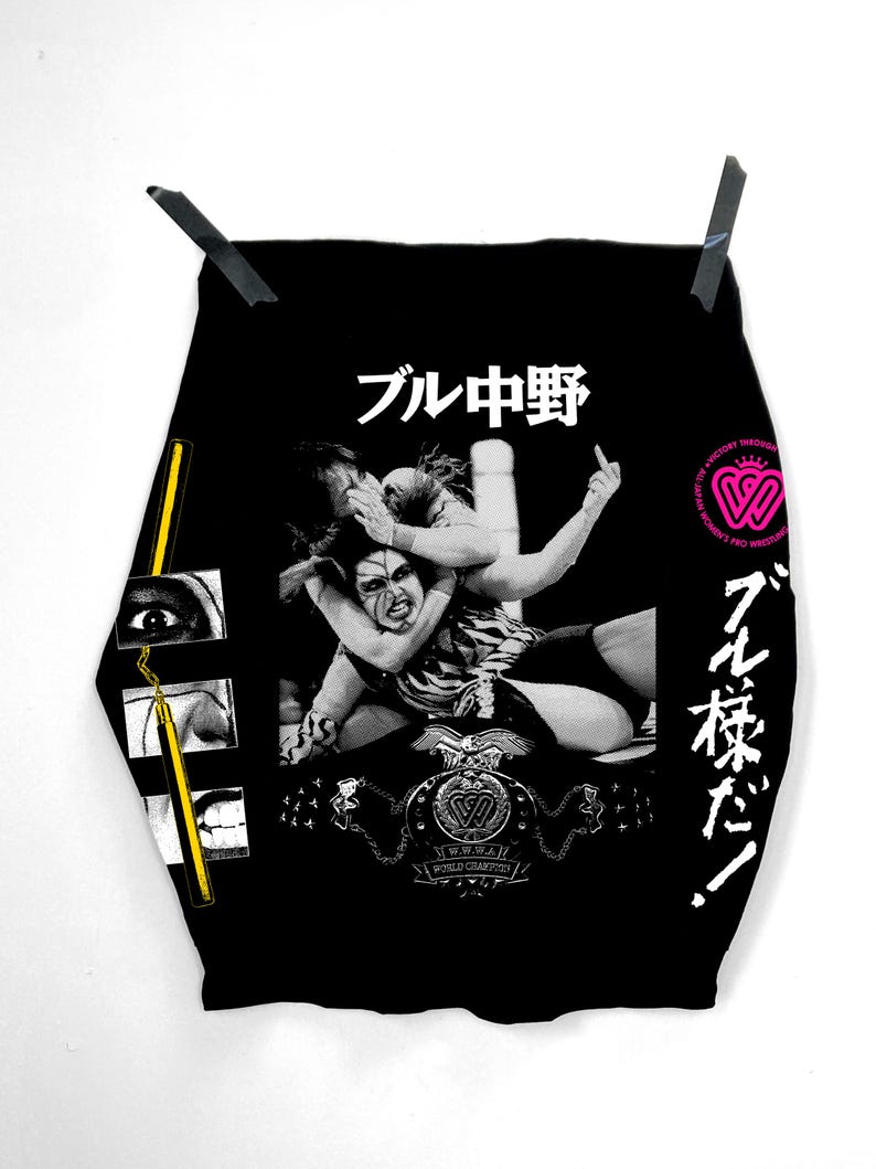 May include: Black graphic tee with a grayscale wrestling illustration. The shirt features Japanese text and a logo for All Japan Women's Pro-Wrestling. The tee is suspended against a white backdrop.