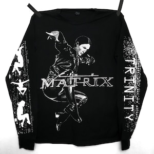 Puede incluir: Camiseta negra de manga larga con un gráfico de Trinity de The Matrix. La imagen muestra a Trinity en una pose dinámica, con la palabra "MATRIX" en el pecho. La manga derecha tiene la palabra "TRINITY" y la frase "Your men are already..." impresa.