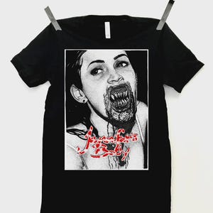 Puede incluir: Camiseta negra con un gráfico en blanco y negro de una mujer con dientes afilados y sangre. El texto "Jennifer's Body" está en rojo, con una escritura estilizada. La camiseta está colgada de un tendedero.