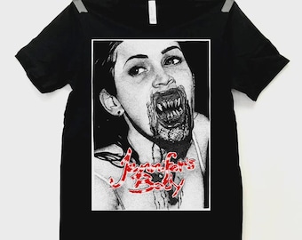 May include: Black t-shirt featuring a black and white graphic of a woman with sharp teeth and blood. The text "Jennifer's Body" is in red, stylized script. The shirt is hanging from a clothesline.
