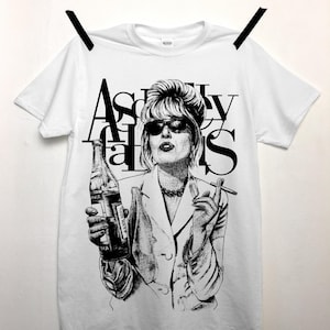 May include: White t-shirt featuring a black and white illustration of a woman with sunglasses, holding a bottle and a cigarette. The text "Absolutely Fabulous" is printed above the illustration in a bold, stylized font. The shirt is hanging on a clothesline.