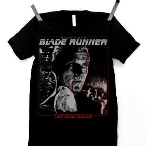 May include: Black t-shirt featuring a graphic from the film "Blade Runner." The design includes the film's title, a portrait of a character, and the phrase "Like Tears in Rain." The shirt is hanging from a clothesline.