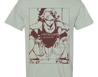 May include: Sage green t-shirt featuring a brown graphic of a character from Metal Gear Solid. The character is in a crouched position, wearing a tank top with the text "METAL GEAR SOLID".