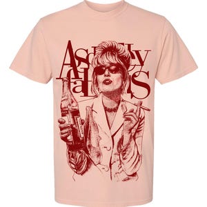 Puede incluir: Camiseta rosa claro con una ilustración burdeos de una mujer con gafas de sol, sosteniendo una botella y un cigarrillo. El texto "Absolutely Fabulous" está impreso en burdeos encima de la ilustración.