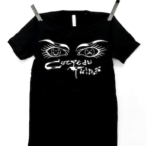 May include: Black t-shirt featuring a white graphic of two eyes, one smiling and one sad, above the text "CozYead Twins". The shirt is suspended by two silver clips on a clothesline.