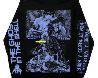 May include: Black long-sleeve shirt featuring a graphic from "The Ghost in the Shell." The design includes a detailed illustration of characters and the text "THE GHOST IN THE SHELL" and "FOUND A VOICE, NOW IT NEEDS A BODY."