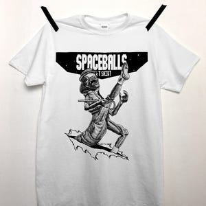 May include: White t-shirt with a black and white graphic of a creature from the movie Spaceballs. The text "SPACEBALLS T SHIRT" is printed above the graphic. The shirt is hanging on a clothesline.