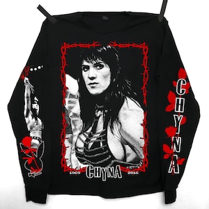 May include: Black long-sleeve shirt with a white image of a woman with long black hair. The image is framed with barbed wire and the text "CHYNA 1969 2016". The sleeves have red roses and the text "CHYNA" in red.