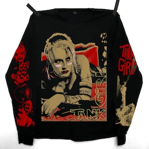 Op de afbeelding: Zwart shirt met lange mouwen met een afbeelding van een vrouw met blond haar en roze accenten, met de woorden "TANK GIRL" in rood en goud. De mouwen hebben extra rode en gouden afbeeldingen.