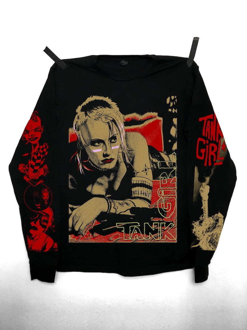 Peut inclure: T-shirt noir &agrave; manches longues avec un graphisme d'une femme aux cheveux blonds et aux accents roses, avec les mots "TANK GIRL" en rouge et or. Les manches ont des graphismes rouges et or suppl&eacute;mentaires.