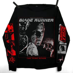 May include: Black long-sleeve shirt featuring a graphic from the film "Blade Runner." The design includes the film's title, a portrait, and the phrase "Like Tears in Rain." Red accents are on the sleeves.