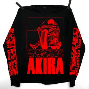 Puede incluir: Camiseta negra de manga larga con un gráfico rojo de una persona en una motocicleta y la palabra "AKIRA" en letras rojas grandes. Las mangas tienen texto rojo y gráficos de pastillas. La camiseta está colgada de pinzas negras.