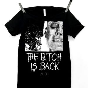 May include: Black t-shirt with a white graphic print of a woman's face and a creature with the text "THE BITCH IS BACK" and "ALIENS" below.