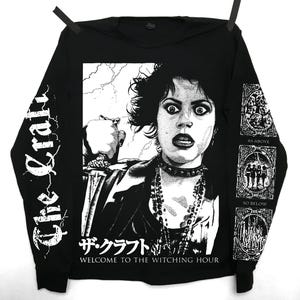 Puede incluir: Camiseta negra de manga larga con un gráfico en blanco y negro de una mujer sosteniendo una daga. El texto "The Craft" está en la manga izquierda. El gráfico incluye texto japonés y la frase "Welcome to the Witching Hour".