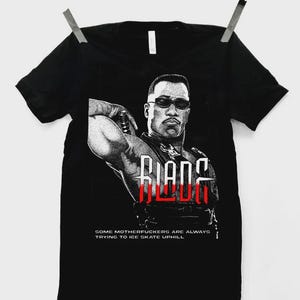 May include: Black t-shirt featuring a graphic of a man in sunglasses with the word "BLADE" in red and white. The text "SOME MOTHERFUCKERS ARE ALWAYS TRYING TO ICE SKATE UPHILL" is below the graphic. The shirt is hanging on a clothesline.