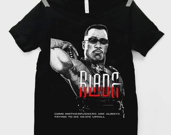May include: Black t-shirt featuring a graphic of a man in sunglasses with the word "BLADE" in red and white. The text "SOME MOTHERFUCKERS ARE ALWAYS TRYING TO ICE SKATE UPHILL" is below the graphic. The shirt is hanging on a clothesline.