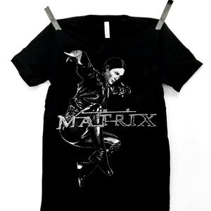 May include: Black t-shirt featuring a graphic of a person in a dynamic pose, with the word "MATRIX" in a stylized font. The image is in black and white. The shirt is hanging from a clothesline.